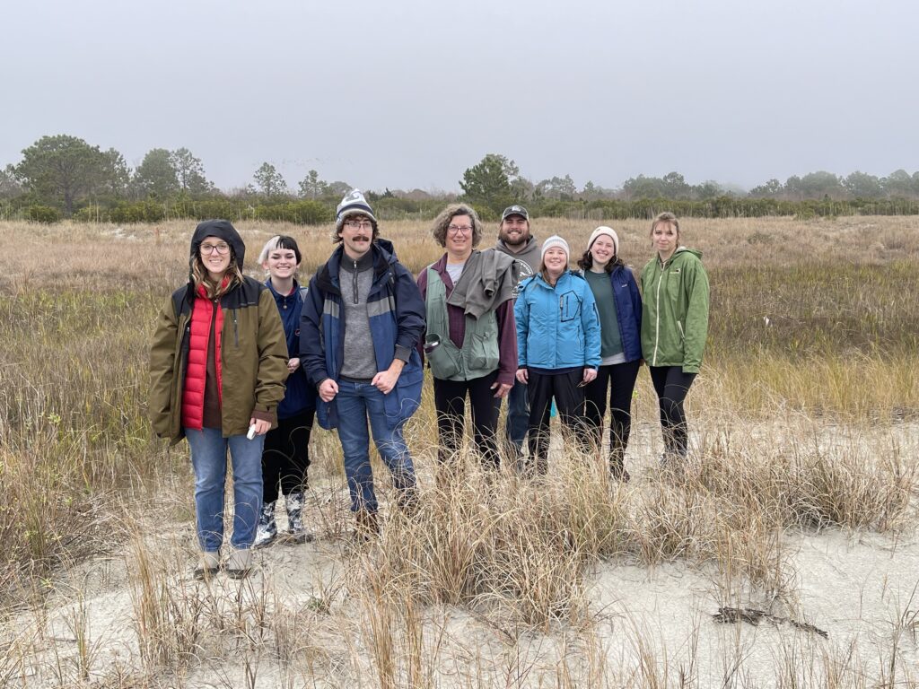 Eight individuals stand on a sandy dune amidst a plethora of grasses. The sky is overcast and light gray and all the individuals are wearing jackets to keep warm. Behind them the grasses grow thicker and more numerous until they give way to shrubs and trees.