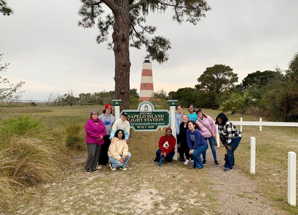 A group of people gathered in front of the Sapelo Island Light Station sign smiling for the camera with the lighthouse in the background