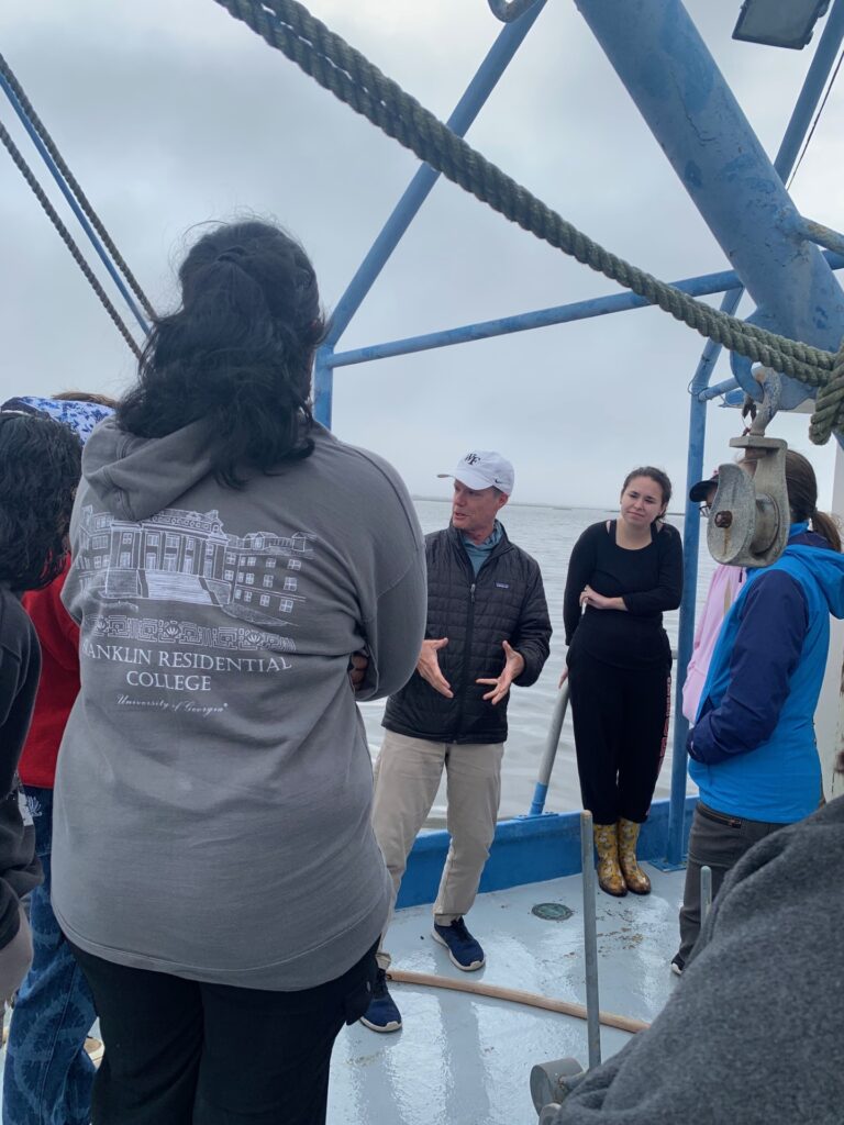 Several people stand on the deck of a trawler facing Assistant Director for Academics Tom Hancock as he speaks to the group. The group is bundled up against the wind and the back of the sweater of the person closest to the camera says "Franklin Residential College"