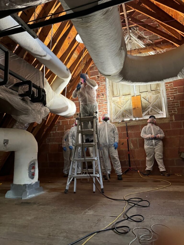 Six people stand in an old dairy barn attic in full protective equipment of white jumpsuits, blue gloves, and face shields with air filters. One figure is standing partway up a ladder in the middle of the attic, using a special spray applicator to disperse the requisite chemicals to fight termites. Large air ducts elbow through the space and the brick wall with wooden window behind the standing figures is lit by a portable light on a stand, illuminating the scene.