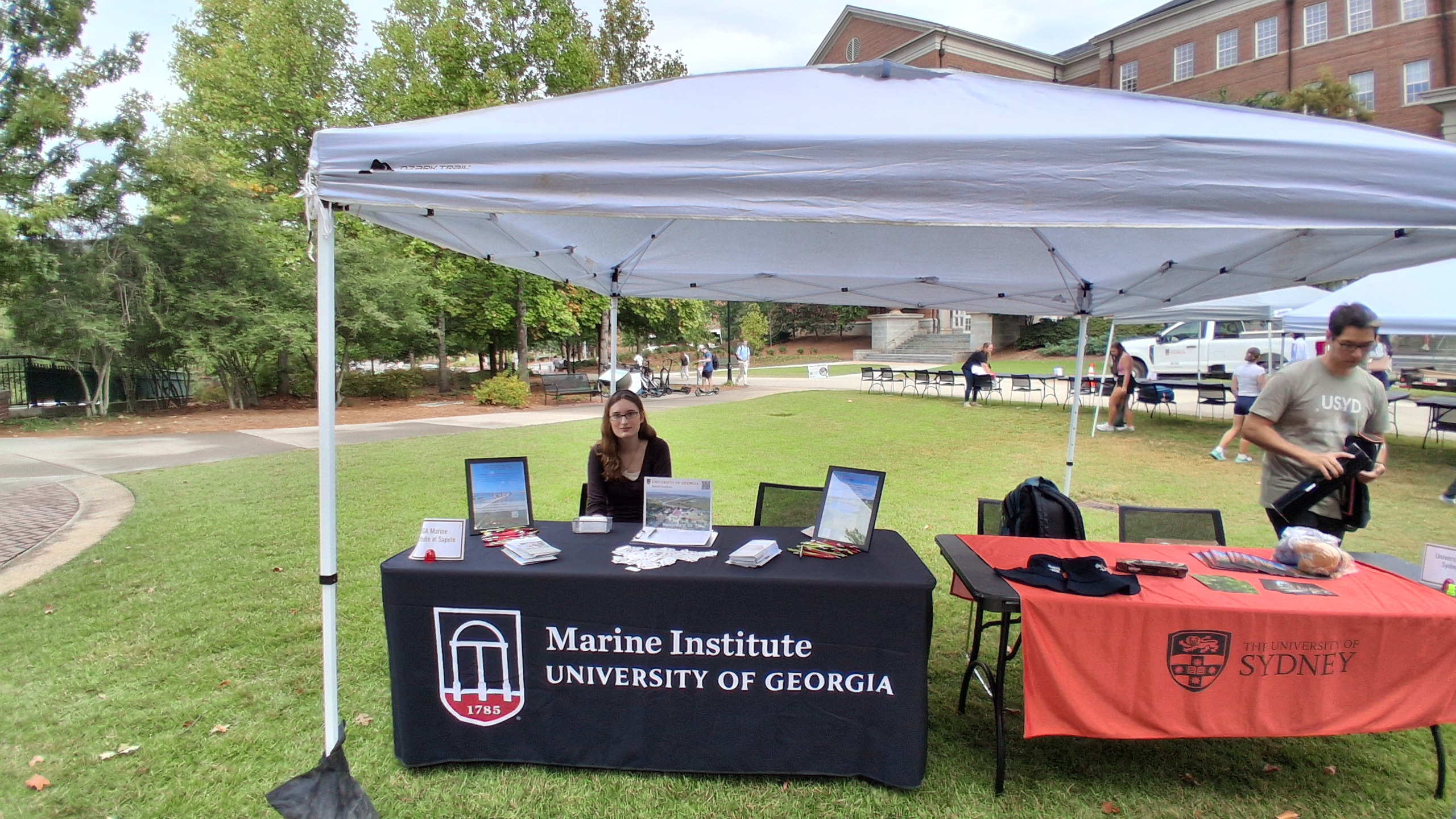 Student sitting outside under a tent at the Study Away Fair, behind a table covered in a black tablecloth emblazoned with the UGA Arches logo and the words "Marine Institute University of Georgia". There are photos, flyers, pens, and stickers available on the table for interested students.