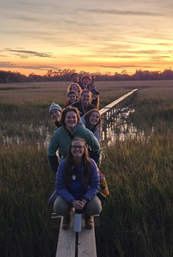 Eleven students smiling on the Teal Boardwalk over the marsh at sunset, arranged poking their heads out from behind each other, a beautiful orange sky behind them