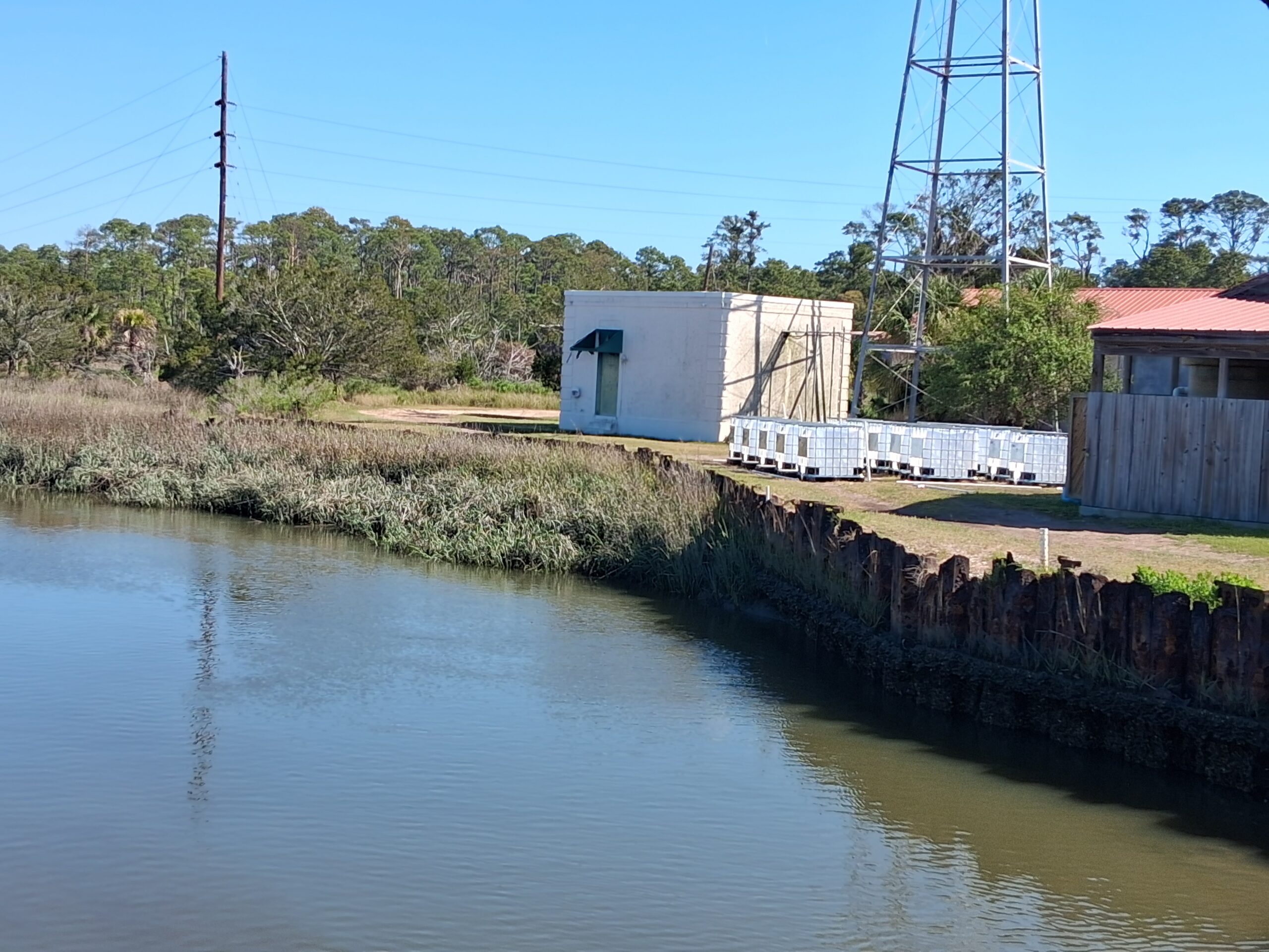 A small utility building stands near a waterway, serving as a UGAMI mesocosm proof of concept; stacked white containers and a metal tower sit nearby, with trees and power lines in the background.
